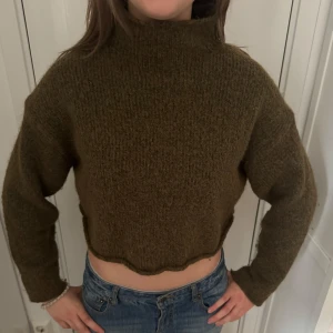 Olivgrön croppad stickad tröja - Säljer en olivgrön croppad stickad tröja med hög hals. Tröjan har lång ärm och en rå, vågig kant nertill för en trendig look. Perfekt till jeans eller kjol för en avslappnad stil.