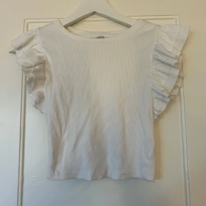 Vit ribbad topp med volangärmar från Zara - Säljer en vit ribbad topp från Zara med korta volangärmar. Toppen har en rund halsringning och är croppad i modellen.