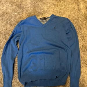 Blå v-ringad tröja från Ralph Lauren - Säljer en klassisk blå v-ringad tröja från Ralph Lauren med broderad logga på bröstet. Tröjan har långa ärmar och ribbade muddar vid ärmslut och nederkant. Perfekt för lager-på-lager-stil.