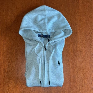 Grå hoodie från Polo Ralph Lauren - Snygg ljusgrå hoodie från Polo Ralph Lauren med dragkedja och justerbar huva. Klassisk broderad logga på bröstet och två fickor framtill. Perfekt för en avslappnad och stilren look.