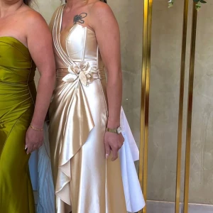 Champangefärgad satinklänning - Guld/beige satinklänning med släp och slit. Använd 2 gånger och i mycket bra skick. Står att den är i size 40 men passar mer en M