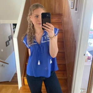 Blå blus med broderade detaljer - Säljer en blå blus med vita broderade detaljer vid halsringningen och axlarna. Blusen har korta volangärmar och dekorativa tofsar framtill. Blusen är aldrig använd.
