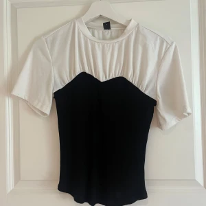 Svart och vit topp med illusion av bustier - Säljer en snygg topp med korta ärmar i vit och svart. Toppen har en cool design där den svarta delen ser ut som en bustier över en vit t-shirt. Perfekt till jeans eller kjol för en trendig look.