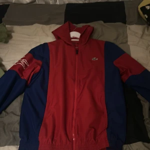 Röd och blå vindjacka från Lacoste Sport - Säljer en snygg vindjacka från Lacoste Sport i rött och blått. Jackan har dragkedja framtill, huva och en broderad Lacoste-logga på bröstet. Perfekt för dig som vill ha en sportig och stilren look.