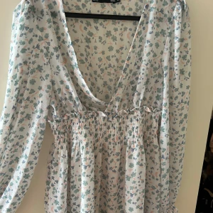 Blommig v-ringad klänning från Lager-157 - Söt blommig klänning från Lager-157 med v-ringning och långa ärmar. Klänningen har smock i midjan och ett luftigt, lätt material. Perfekt för varma dagar och har ett romantiskt mönster i pastellfärger.