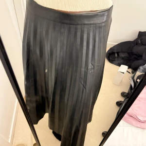 Svart långkjol i skinnimitation - Säljer en svart långkjol med diskreta vertikala ränder i skinnimitation. Kjolen har en hög midja och ett stilrent, klassiskt snitt som ger en snygg siluett. Perfekt för dig som gillar en edgy och trendig look.