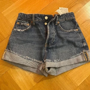 Zara shorts - Säljer ett par blå jeansshorts från Zara med råa fransade kanter och uppvikta benslut. Klassisk femficksmodell med knappgylf och bälteshällor. Perfekta för varma dagar och enkel att matcha med olika toppar.