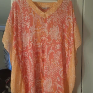 Mönstrad kaftan från KappAhl - Säljer en luftig och färgglad kaftan från KappAhl i orange och vitt med blommigt mönster. Plagget har v-ringning och är tillverkat i ett lätt och syntetiskt material, perfekt för varma dagar.