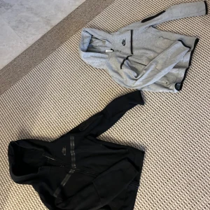 Nike Tech Fleece ziphoods i svart och grå - Två Nike Tech Fleece ziphoods säljes, en i svart och en i ljusgrå. Båda har huva, dragkedja och svarta detaljer längs ärmar och bröst. Snygg sportig look med smal passform och klassisk Nike-logga på bröstet.