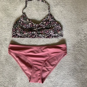 Blommig bikini med rosa underdel - Supersöt bikini med blommigt mönstrad topp i svart, rosa och vitt samt matchande enfärgad rosa underdel. Toppen har smala axelband och lätt rynk framtill. Perfekt för stranden eller poolen i sommar!