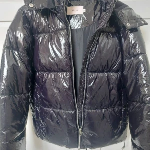 Svart glansig pufferjacka från Nelly - Svart pufferjacka från Nelly med glansig finish och quiltad design. Jackan har huva, dragkedja framtill och dragsko nertill. Perfekt för kalla dagar och riktigt snygg till jeans eller mjukisbyxor.