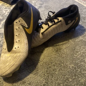 Nike phantom academy svart/guld EU 40 - Snygga fotbollsskor från Nike i svart och vitt med guldiga detaljer och klassisk swoosh. Skorna har snörning och mönstrad ovandel för extra grepp. Yttersulan har dobbar för bra fäste på planen. Perfekta för dig som vill sticka ut på matchen!. Ser smutsig ut pågrund av spel på konstgräs (det går att ta bort). Sko kartongen följer INTE MED.