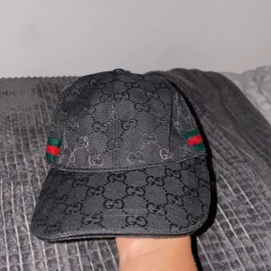Svart Gucci keps med GG-mönster - Snygg svart keps från Gucci med klassiskt GG-monogram över hela kepsen och ikoniska gröna och röda ränder på sidan. Justerbar rem baktill i skinn och diskret logotyp på insidan. Perfekt accessoar för att lyfta din outfit.