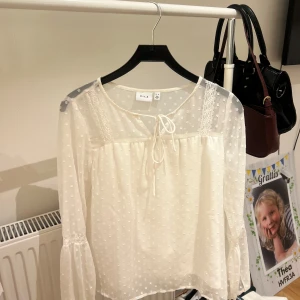 Vit prickig blus från Vila - Supersöt vit blus från Vila med transparenta, långärmade ärmar och diskret prickigt mönster. Blusen har knytning vid halsen, spetsdetaljer på axlarna och volang vid ärmsluten. 