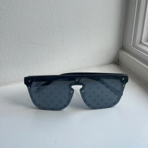 Louis Vuitton blå monogram solglasögon - Snygga solglasögon från Louis Vuitton med spegellins och ikoniskt monogrammönster över hela glaset. Sköldformad modell med bred båge och silvriga nitar vid kanterna. Perfekt för dig som vill sticka ut med en lyxig vibe.