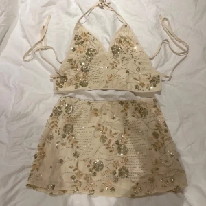 Beige paljett-topp med blommor - Supersöt croppad linnetopp i beige med blommigt mönster och massor av glittriga paljetter. Toppen har smala knytband i nacken och ryggen, v-ringad front. Sjukt snyggt set och perfekt för festivaler i sommar💛