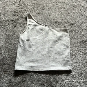 Vit oneshoulder topp från Gina Tricot - Säljer en stilren vit oneshoulder topp från Gina Tricot i ribbad bomull. Toppen är ärmlös med asymmetrisk axel och har en tight passform som ger en modern vibe. Perfekt att matcha med jeans eller kjol för en trendig look.