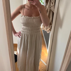 Beige långklänning med smock - Supersöt beige långklänning med smala axelband och smockad överdel. Klänningen har en luftig och flowy passform, perfekt för varma dagar. Den är ärmlös och har en enkel, stilren look som är lätt att matcha med accessoarer.