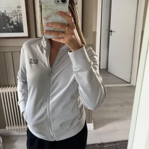Vit zip-jacka från Emporio Armani - Snygg vit zip-jacka från Emporio Armani med EA7-logga på bröstet och längs ryggen. Liten i storleken. 