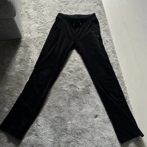 Svarta velourbyxor Juicy Couture XS - Svarta mjukisbyxor från Juicy Couture i velour med glittrande strasstenar på fickan och stor 'Juicy'-text baktill. Byxorna har dragsko i midjan och klassisk rak passform. Perfekta för en chill och trendig look.