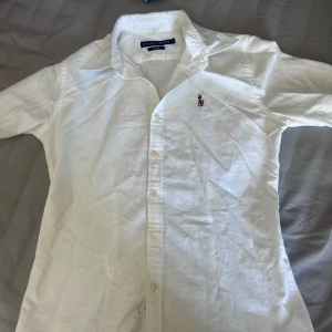 Vit skjorta från Ralph Lauren - Klassisk vit skjorta från Ralph Lauren med broderad logga på bröstet. Skjortan har lång ärm, knappar framtill och en snygg krage. Perfekt för en clean och stilren look.