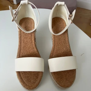 Vita kilklackade sandaler med repdetalj - Snygga sandaler med vit rem över tårna och runt ankeln, justerbart spänne i guld och kilklack täckt av flätat rep. Sulan är i kork och ovandelen i syntetmaterial. Perfekta för sommarens alla tillfällen.