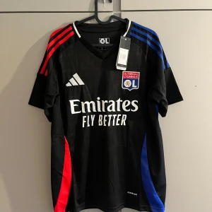 Olympique Lyonnais svart matchtröja Adidas - Snygg svart fotbollströja från Adidas för Olympique Lyonnais med röda och blå detaljer på axlar och sidor. Tröjan har klubbmärke på bröstet, Adidas-logga och Emirates-tryck framtill. Tillverkad i lätt och ventilerande polyester.