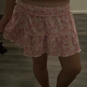 Rosa blommig volangkjol - Supersöt kort kjol med blommigt mönster i rosa och vitt. Kjolen har smock i midjan och volang nertill som ger en flowy känsla. Perfekt för varma dagar och lätt att matcha med en enkel topp.
