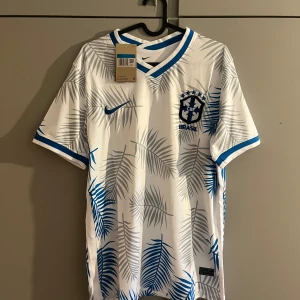 Vit Brasil Nike fotbollströja palmblad - Snygg vit Brasilien fotbollströja från Nike med blå detaljer och palmbladsmönster i grått och blått. Tröjan har korta ärmar, V-ringad krage med blå kant och broderat CBF-märke på bröstet. Tillverkad i lätt och ventilerande material, perfekt för match eller träning.