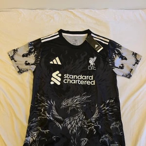 Liverpool x Adidas svart fotbollströja S - Liverpool fotbollströja från Adidas i svart med gråa detaljer och ett mäktigt drakmönster över hela tröjan. Kortärmad modell med vita Adidas-ränder på axlarna och klubbmärke samt sponsortryck på bröstet. Tillverkad i lätt och ventilerande material.