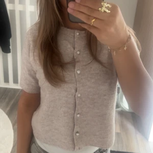 Beige kofta med pärlknappar - Supersöt beige kofta med korta ärmar och pärlknappar framtill. Den är stickad och har en mjuk, mysig känsla som passar perfekt till lager-på-lager. Enkel att styla med både jeans och kjol för en chill look.