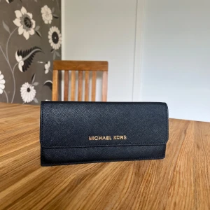 Svart plånbok från Michael Kors - Stilren svart plånbok från Michael Kors i skinn med guldfärgad logga framtill. Perfekt för dig som vill ha en snygg och enkel accessoar.