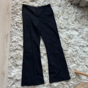 Svarta kostymbyxor från Gina Tricot - Snygga svarta kostymbyxor från Gina Tricot med raka pressveck och bootcut-silhuett. Byxorna har hög midja och är tillverkade i ett mjukt material som ger en clean och stilren look. De är i fint skick, men blivit noppriga runt dragkedjan.