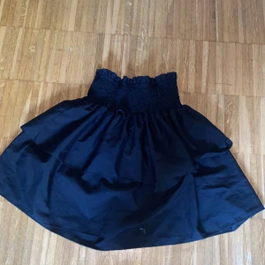 Svart volangkjol med smock - Supersöt svart kortkjol med bred smock i midjan och dubbla volanger. Kjolen har en luftig känsla och passar perfekt till sneakers eller boots. Enkel att styla och ger en cool vibe till din outfit.