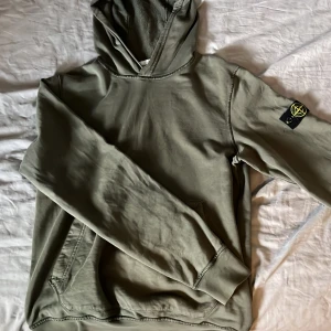 Olivgrön hoodie från Stone Island - Snygg olivgrön hoodie från Stone Island med klassisk patch på ärmen. Tröjan har huva, känguruficka och långa ärmar. Perfekt för dig som gillar streetwear och vill ha en stilren look med ikonisk logga. Köpt på NK original pris 2000kr. Inga defekter.