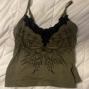 Olivgrön linnetopp med spetsdetalj - Snygg olivgrön linnetopp med smala axelband och svart spets längs v-ringningen. Framsidan har ett coolt svart tryck med fjärilsmotiv och grafiska detaljer. Toppen är figurnära och har justerbara axelband för perfekt passform.