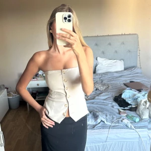 Beige korsett-topp med knappar - Säljer en beige axelbandslös korsett-topp med snygg knäppning framtill. Toppen har en figurnära passform och markerad midja som ger en cool siluett. Perfekt att styla med både kjol och jeans för en trendig look.