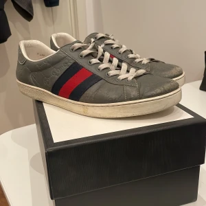 Gucci sneakers med röd och marinblå rand - Snygga sneakers från Gucci i grått skinn med klassiska ränder i rött och marinblått på sidan. Skorna har snörning, vit sula och präglade GG-loggor i lädret. Perfekta för dig som vill ha en lyxig och stilren look.