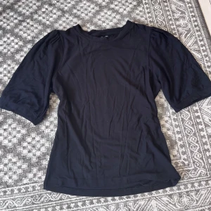 Svart topp med puffärm från 157 - Svart topp från 157 The Black Label Collection med puffiga korta ärmar och rund halsringning. Toppens material känns mjukt och lätt, perfekt för en clean och stilren look. Passformen är normal och modellen är enkel men trendig. Bra skick och nytvättad 💓