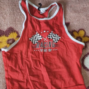 Röd linnetopp från Divided XS - Cool röd linnetopp från Divided med vita kanter och tryck med racingflaggor och texten 'Racing to the top' framtill. Perfekt för dig som gillar sportig stil och vill sticka ut. Tunt och mjukt bomullsmaterial, ärmlös och med brottarrygg.