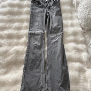 Grå bootcut jeans med låg midja - Snygga grå jeans med bootcut-modell och hög midja. Jeansen har klassiska fem fickor och är tillverkade i mjukt denimtyg. Perfekta för dig som gillar en retrovibe och vill ha ett par jeans som sticker ut lite extra.