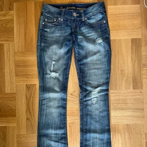 Blå slitna jeans från Guess - Snygga lågmidjade blå jeans från Guess med slitna detaljer och raka ben. 