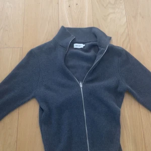 Mörkgrå ribbad kofta från East West - Snygg mörkgrå ribbad kofta från East West med hel dragkedja framtill och hög krage. Perfekt för lager-på-lager och enkel att matcha. Långärmad och i stickad bomullsmix som känns både mjuk och stilren. Storlek M men passar som S pris går att diskutera 