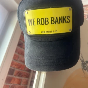 Svart keps John Hatter & Co "We Rob Banks" - Cool svart keps från John Hatter & Co med mesh på sidorna och bak. Framtill sitter en gul skylt med texten "WE ROB BANKS" som ger kepsen en edgy vibe. Justerbar passform och böjd skärm. Perfekt statement-piece för dig som vill sticka ut.