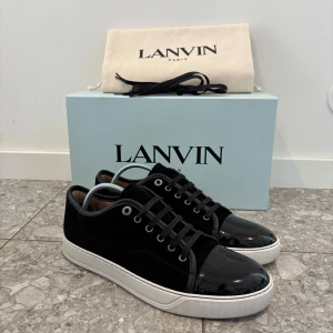 Lanvin Cap Toe - Snygga svarta sneakers från Lanvin med vit platt sula och glansig lackad tå. Skorna har snörning och är tillverkade i en mix av mocka och skinn, vilket ger en lyxig känsla. Perfekta för dig som gillar stilrena och exklusiva sneakers. Skorna är sparsamt använda ett par gånger. Storleken är UK 9. Köpta på NK.