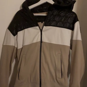 Moose Knuckles beige och svart jacka - Snygg jacka från Moose Knuckles i beige, vitt och svart med coolt quiltat parti upptill. Jackan har huva, dragkedja framtill och två fickor. Märkespatch på ärmen och stilren design som sticker ut. Perfekt för dig som gillar streetwear och vill ha något unikt.