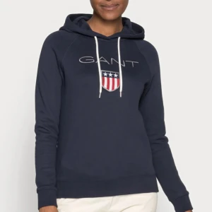 Mörkblå hoodie från GANT  - Säljer en mörkblå hoodie från GANT med broderad logga på bröstet. Tröjan har huva med vita snören och långa ärmar. 