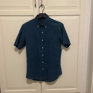 Blå kortärmad skjorta Ralph Lauren - Snygg blå skjorta från Ralph Lauren med korta ärmar och klassisk button-down krage. Skjortan har knappar hela vägen och en diskret broderad logga på bröstet. Perfekt för en stilren och avslappnad look.