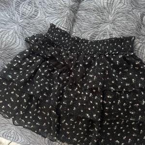 Svart blommig volangkjol - Supersöt svart kjol med små blommor och flera volanger. Kjolen har bred smock i midjan för en bekväm passform och är gjord i ett lätt, luftigt material. Perfekt för sommaren.   Kjolen är dessutom ny och oanvänd.