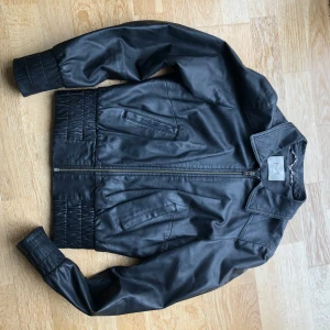 Svart skinnjacka H&M x Madonna strl S - Snygg svart skinnjacka från H&M i klassisk bombermodell. Jackan har dragkedja framtill, två fickor och breda ribbade muddar vid ärmslut och midja. Perfekt för dig som vill ha en stilren och edgy look till vardags.